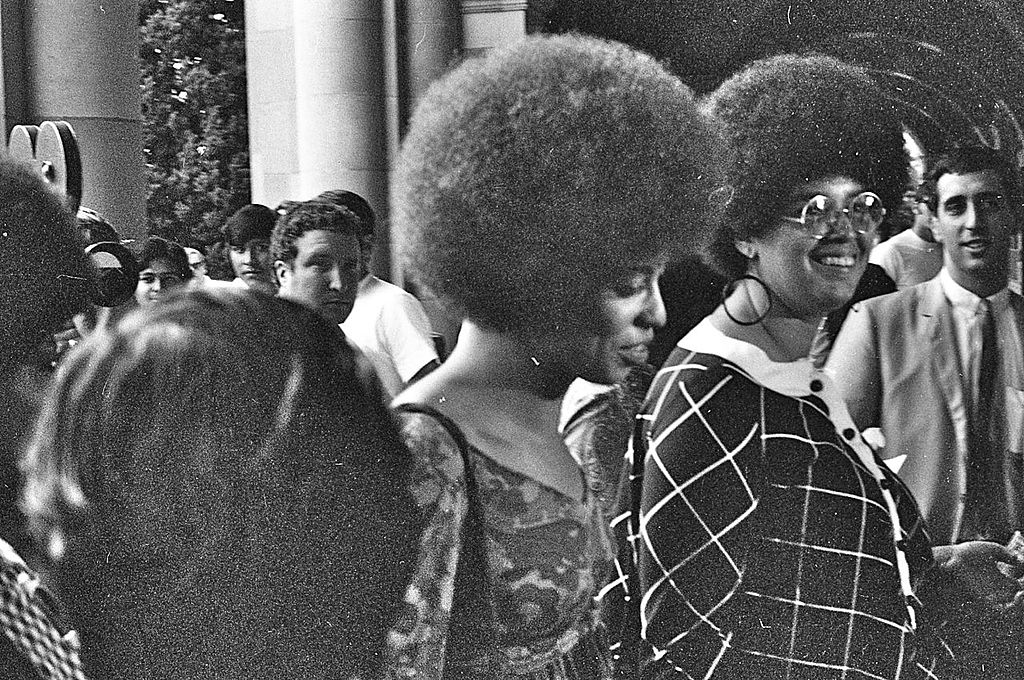 Angela Davis walking, surrounded by a crowd of people, followed by a film crew.