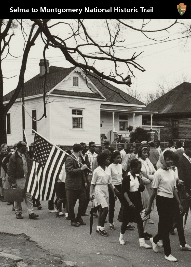 Young women and young men marching with American flag. Residential street with homes in background.